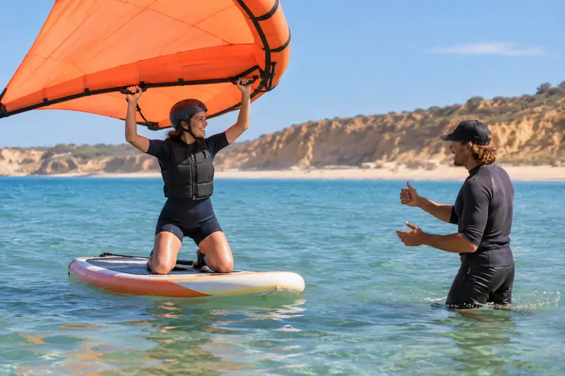 Una alumna de wing foil navega de rodillas sobre una tabla grande con el ala en las manos, en agua tranquila